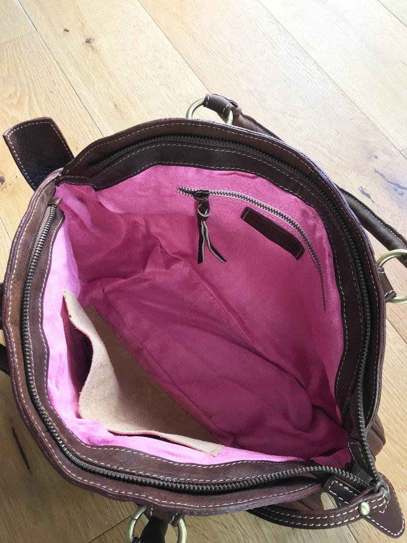 May include: Brown leather handbag with a pink fabric lining and a zippered pocket. The bag has a top handle and a zipper closure.