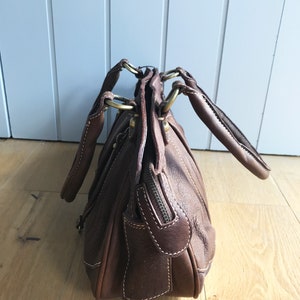 May include: A brown leather handbag with gold hardware and a zipper closure. The bag has two top handles and a shoulder strap.