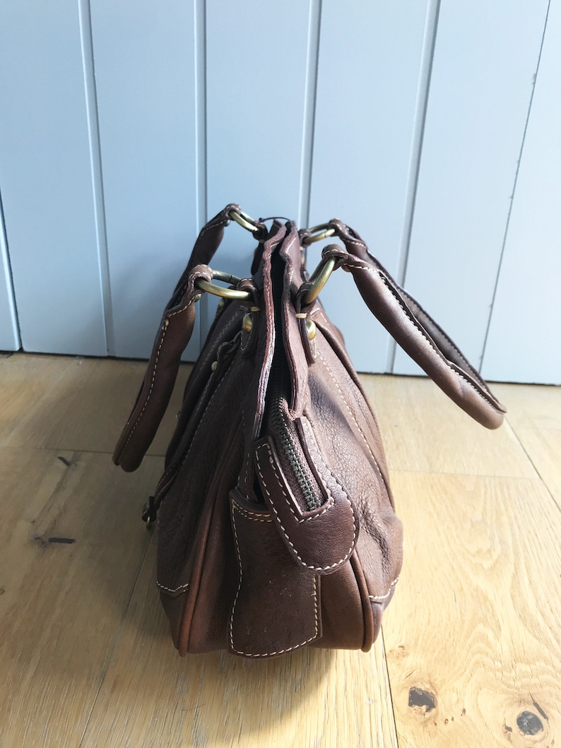 May include: A brown leather handbag with gold hardware and a zipper closure. The bag has two top handles and a shoulder strap.