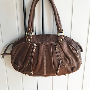 May include: A brown leather handbag with gold-toned hardware and a top zipper closure. The bag has two top handles and a detachable strap.