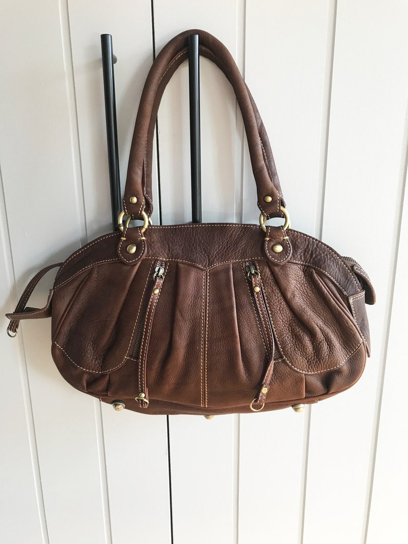 May include: A brown leather handbag with gold-toned hardware and a top zipper closure. The bag has two top handles and a detachable strap.