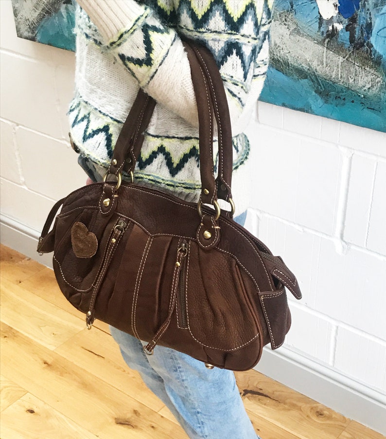 May include: A brown leather handbag with a heart-shaped charm and gold-toned hardware. The bag has a top handle and a zipper closure.