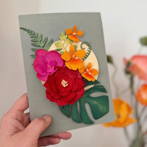 Op de afbeelding: Handgemaakte wenskaart met een bloemenontwerp. De kaart heeft een rode roos, roze orchidee en oranje bloemen, samen met groene bladeren. De bloemen zijn gerangschikt op een lichtgroene achtergrond.