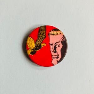 May include: A red round button featuring a black and yellow eagle in flight on the left and a man's face on the right. The man has blonde hair and a serious expression.