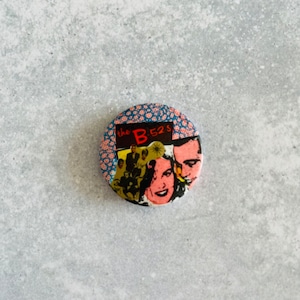 May include: A circular badge with a collage design. The top section has a blue and red patterned fabric design. The bottom section features a band photo and the text "the B-52's". The badge has a shiny surface.