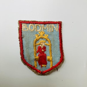 Peut inclure: Écusson brodé avec le mot "BODMIN" en fil d'or. Le patch présente un fond bleu clair, une arche dorée et une figure rouge. Il est bordé de coutures rouges et marron. Un emblème de style vintage.
