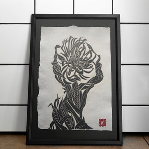 Op de afbeelding: Een zwart-witte linosnede in een zwarte lijst met een gestileerd silhouet van een hoofd gevormd door ingewikkelde bloem- en bladpatronen. Het kunstwerk staat op getextureerd papier met gescheurde randen en bevat een kleine rode vierkante stempel met een witte letter K.