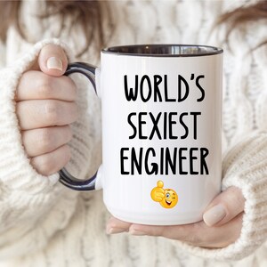 Könnte beinhalten: Weißer Keramikbecher mit schwarzem Innenraum und Henkel. Der Becher zeigt den Text "WORLD'S SEXIEST ENGINEER" in Schwarz, zusammen mit einem Daumen hoch Emoji. Der Becher wird von einer Person gehalten, die einen weißen Pullover trägt.