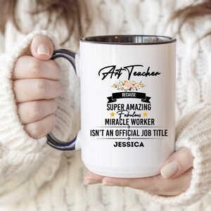 Peut inclure: Mug en céramique blanche avec intérieur et anse noirs. Le mug affiche le texte "Art Teacher" au-dessus d'un motif floral, suivi de "BECAUSE SUPER AMAZING FABULOUS MIRACLE WORKER ISN'T AN OFFICIAL JOB TITLE JESSICA."
