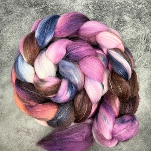 Könnte beinhalten: Ein farbenfroher Zopf aus Wollvlies in Rosa-, Lila-, Blau- und Brauntönen. Die Faser ist kreisförmig angeordnet und hat ein weiches, strukturiertes Aussehen. Das Vlies eignet sich zum Spinnen, Filzen oder für andere Faserprojekte.