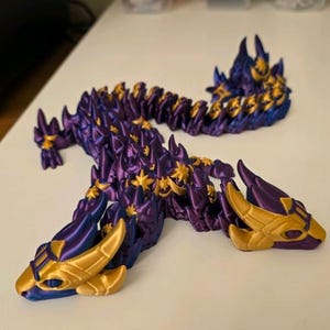 May include: A purple and gold articulated dragon toy. The dragon is made of plastic and has a segmented body, allowing for flexible movement. The dragon's head and body feature gold accents, with a gradient of blue and purple.