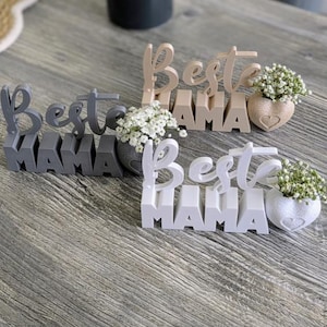 May include: Three wooden signs saying "Best Mama" in grey, beige, and white. Each sign has a small vase with white flowers. The vases are heart-shaped or round. The signs are displayed on a wooden surface.