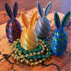 May include: Four colorful Easter egg decorations with bunny ears. The eggs are in shades of red, gold, silver, and blue, with a striped design. They are arranged on a wooden surface with gold and green beads.