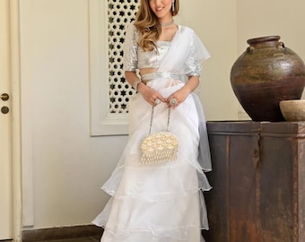 May include: A white layered saree with a silver sequined blouse and a matching belt. The outfit includes a small, beaded clutch. The saree has a tiered design, and the blouse has short sleeves. The overall look is elegant and formal.