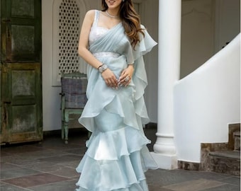 May include: A light blue saree with a layered, ruffled skirt and a matching draped shawl. The top is a silver sequined bodice. The saree is worn with a silver belt and jewelry. The background includes a white column and a dark green door.