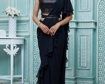 Puede incluir: Un vestido de noche negro con un corpiño de lentejuelas y una falda con volantes. El vestido presenta un diseño de un solo hombro y un cinturón a juego. La modelo lleva joyas de plata. El fondo es una pared azul claro con paneles decorativos.