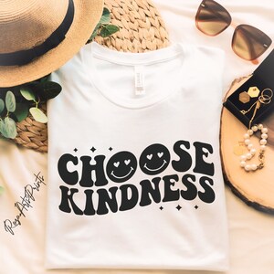 Può includere: Una maglietta bianca con la scritta "CHOOSE KINDNESS" in nero, con un font retrò. Il design include faccine sorridenti con occhi a cuore e dettagli a stella. La maglietta è presentata con occhiali da sole e gioielli.