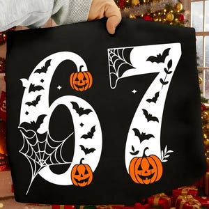 May include: Black Halloween-themed pillow with the numbers 67 in white. The numbers are decorated with bats, spiderwebs, and orange jack-o'-lanterns. The pillow is held by a person.