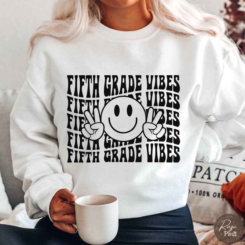 May include: White sweatshirt with black text that reads "FIFTH GRADE VIBES" repeated in a wavy pattern. A black smiley face with peace signs is centered on the sweatshirt.