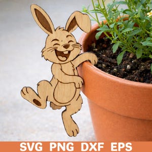 Könnte beinhalten: Eine Holzdekoration eines lächelnden Hasen, der sich an einem Terrakotta-Topf abstützt. Der Hase ist hellbraun mit geätzten Details, und der Topf enthält grüne Pflanzen. Der Text "SVG PNG DXF EPS" befindet sich am unteren Bildrand.