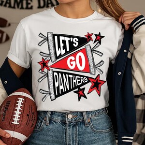 Peut inclure: T-shirt blanc avec un motif graphique comportant le texte "LET'S GO PANTHERS" dans des formes de fanions noir et blanc, avec un fanion rouge au centre. Des &eacute;toiles rouges ornent le texte. Un ballon de football est pr&eacute;sent.