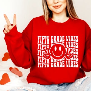 May include: Red sweatshirt with white text that says "Fifth Grade Vibes" repeated multiple times. A white smiley face with peace signs is in the center of the text.