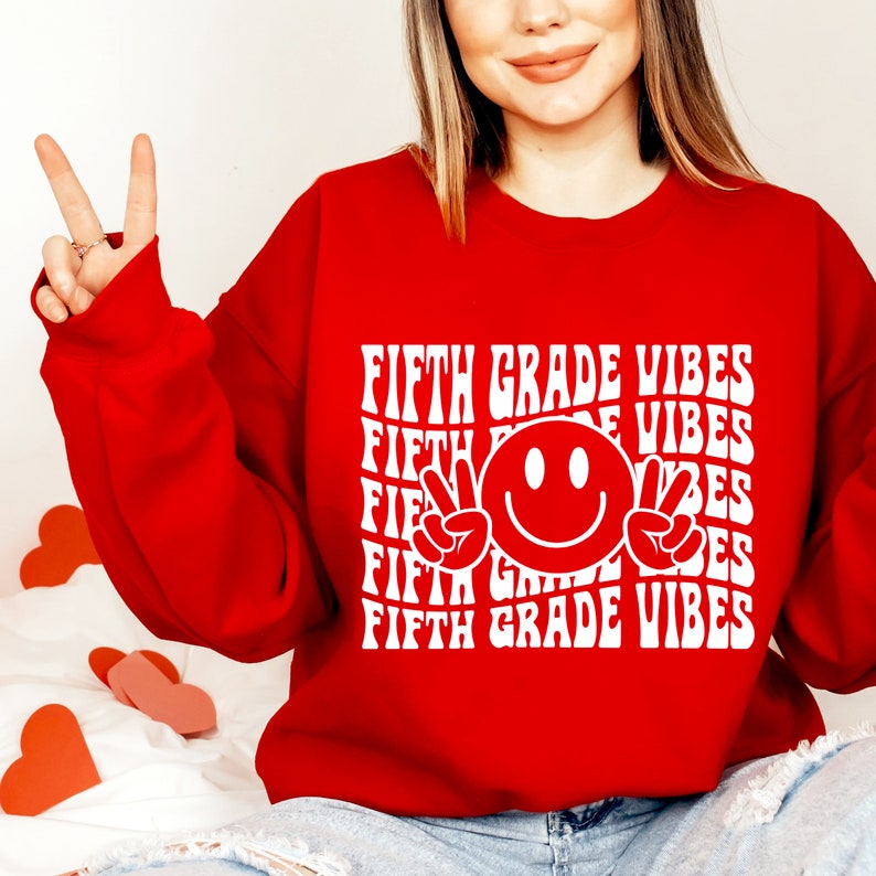 May include: Red sweatshirt with white text that says "Fifth Grade Vibes" repeated multiple times. A white smiley face with peace signs is in the center of the text.