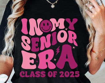 May include: Black t-shirt with pink and purple text that reads "In My Senior Era Class of 2025". The text is in a retro font with a smiley face.