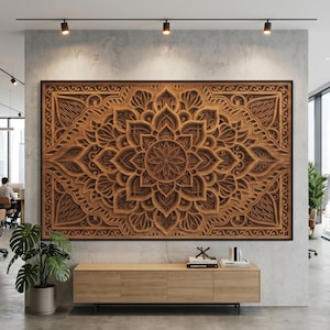Op de afbeelding: Grote, ingewikkeld gesneden houten wandkunst met een mandala-ontwerp. Het kunstwerk is zwart omlijst en hangt aan een grijze muur boven een houten kast. De kantooromgeving omvat planten en railsverlichting.