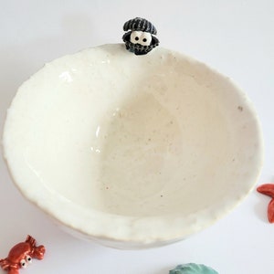 Könnte beinhalten: Eine weiße Keramikschüssel mit einer schwarz-weißen Muschelfigur am Rand. Die Schüssel ist leer und hat eine leicht strukturierte Oberfläche.