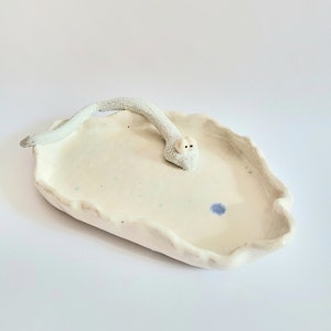 Könnte beinhalten: Handgefertigte Keramikschale mit einer weißen Schlangenfigur. Die Schale ist cremeweiß mit gewelltem Rand und einem blauen Punkt. Die Schlange ist weiß mit strukturierter Oberfläche und kleinen schwarzen Augen. Ein einzigartiges Dekorationsobjekt.