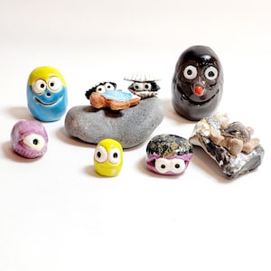 Könnte beinhalten: Eine Sammlung kleiner, handbemalter Keramikfiguren mit Glitz Augen. Die Figuren sind in Form verschiedener Meeresbewohner geformt, darunter eine Schildkröte, eine Muschel und ein Krebs. Es gibt auch eine kleine, runde Figur mit einem schwarz-weißen Gesicht.