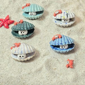 Könnte beinhalten: Sechs Keramikmuscheln mit Glitz Augen und kleinen roten Krabben darauf. Die Muscheln sind in verschiedenen Farben, darunter Blau, Grün und Weiß. Die Muscheln liegen auf einer weißen Sandfläche.