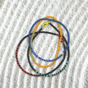 Peut inclure: Plusieurs colliers de perles de différentes couleurs, dont le noir, le rouge, le bleu et le jaune. Chaque collier présente une couleur de fermoir différente, avec un mélange de petites perles colorées. Les colliers sont présentés sur une surface blanche texturée.