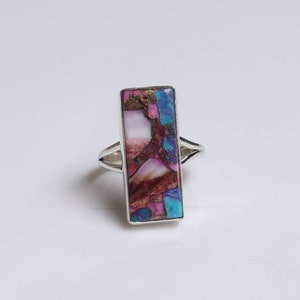 May include: A silver ring with a rectangular, multi-colored stone. The stone is a mix of pink, blue, and brown.