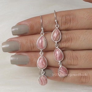 May include: A pair of sterling silver drop earrings featuring three vertically aligned, teardrop-shaped pink rhodochrosite stones in each earring. The stones exhibit natural white and pink banding patterns set in polished silver frames with hook wires.