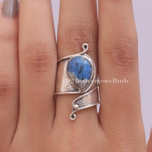 May include: A silver ring featuring a blue and white gemstone. The ring is wrapped around the finger and has a textured finish.