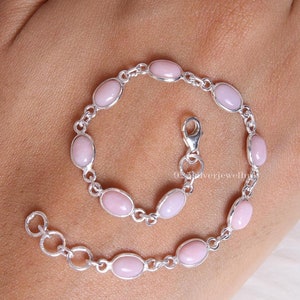 May include: A silver chain bracelet featuring nine smooth, oval-shaped light pink stones set in polished silver bezels. The bracelet includes a lobster claw clasp and an adjustable extender chain with circular links, showcasing a delicate design.