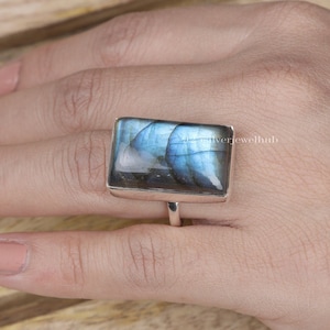 May include: A rectangular labradorite ring with a silver band. The gemstone displays iridescent blue and green hues. The ring is shown on a hand against a light wooden surface. Jewelry.