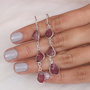 May include: A pair of silver dangle earrings with four pink, rough-cut gemstones on each earring. The earrings are connected by a silver chain.