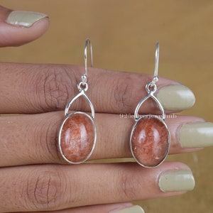 May include: A pair of silver dangle earrings with oval-shaped sunstone gemstones. The earrings have a simple design with a small loop at the top and a delicate drop.