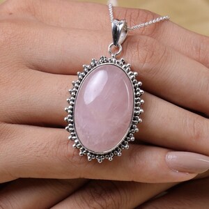 May include: An oval-shaped rose quartz pendant set in a silver frame with a beaded edge detail. The pendant hangs from a delicate silver chain. The stone is smooth, polished, and light pink in color, held within a decorative metallic setting.
