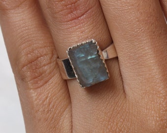 May include: A silver ring with a rectangular, raw-cut labradorite gemstone. The stone displays blue-grey hues with iridescent flashes. The ring is set on a plain, polished silver band. The ring is shown on a person's finger.