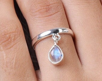May include: A silver ring featuring a smooth, polished band and a single dangling charm. The charm is a teardrop-shaped moonstone with a translucent, iridescent blue glow, securely attached to the band by a small, circular silver jump ring.