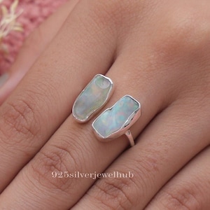 Può includere: Un anello d'argento con due pietre di opale irregolari e chiare. Le pietre hanno una lucentezza perlacea e sono incastonate in una semplice fascia d'argento. L'anello è mostrato su una mano, evidenziando il design e il colore del gioiello.