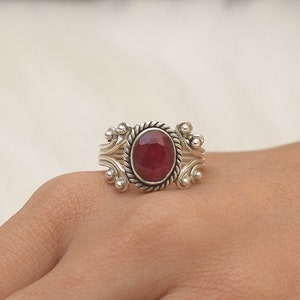 Puede incluir: Un anillo de plata con una piedra preciosa de color rojo oscuro en forma ovalada. La piedra está rodeada por un borde de metal trenzado, flanqueada por adornos de volutas de plata y pequeños detalles esféricos en el anillo.