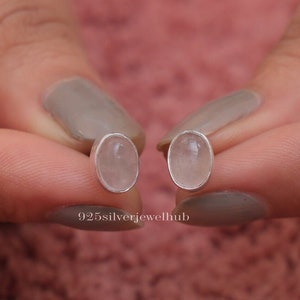Può includere: Un paio di orecchini a lobo di forma ovale con pietre lisce, traslucide e rosa pallido incastonate in montature d'argento lucido. Gli orecchini sono tenuti in mano per mostrare il taglio cabochon. Il testo 925silverjewelhub è visibile sotto.