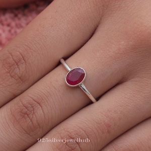 Puede incluir: Un anillo de plata con una piedra ovalada roja. El anillo está en el dedo de una persona.