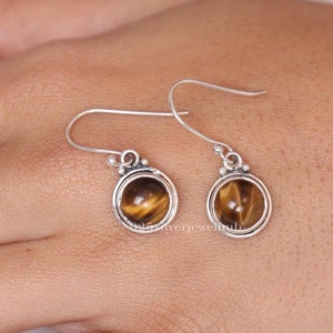 May include: A pair of silver drop earrings with round tiger's eye stones. The earrings have a simple design with a small decorative element above the stone. The tiger's eye stones are a warm brown color with golden stripes.