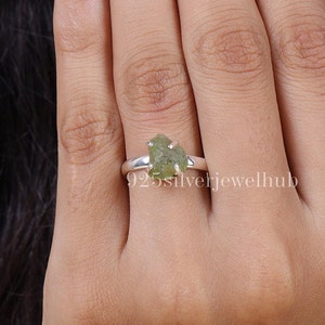 May include: A sterling silver ring featuring a raw, unpolished olive green gemstone held in a four-prong setting. The band is smooth and polished, showcasing a simple metal design that highlights the natural, irregular texture of the stone.